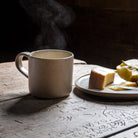Rauchende Steingut Tasse in Beige mit Brotzeit nebendran.